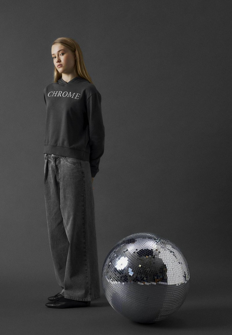 Grå sweatshirt med texten "CHROME", kombinerad med löst sittande mörka jeans. En stor, rund silverfärgad discokula reflekterar ljuset i förgrunden.