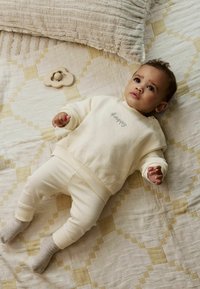 Cremefarvet sweatshirt med "Happy" broderet, parret med ribbede cremeleggings. Ligger på en mønstret quilt med et trælegetøj i nærheden.
