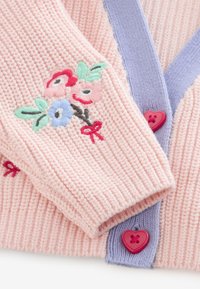 Close-up of a pink knitted sweater sleeve with colorful floral embroidery and purple trim with red heart-shaped buttons.