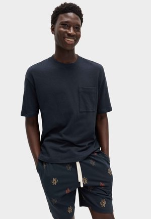 Smilende mand iført en marineblå kortærmet skjorte med brystlomme og marineblå shorts med mønster af små brune og røde kaktusser.