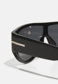 Occhiali da sole neri con una montatura spessa e angolare. Presentano lenti scure e un accento dorato sulle tempie. Texture liscia e lucida.