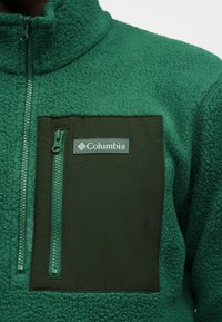 Πράσινο φλις σακάκι Columbia με σκούρο πράσινο τσεπάκι στο στήθος που διαθέτει κάθετο φερμουάρ και λογότυπο Columbia.