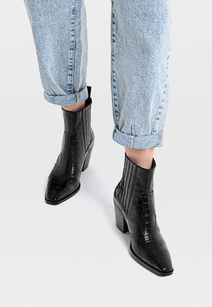 Personne portant un jean bleu clair avec revers et des bottines noires à talons hauts avec une texture en peau de crocodile sur fond blanc.