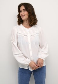 White blouse with a button-up front, pleated texture, lace accents, and puffed sleeves. Paired with blue jeans, featuring a smooth fabric finish.