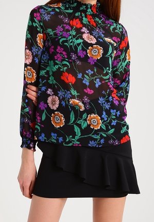 Mujer con una blusa negra transparente floral de manga larga con cuello alto con volantes y una minifalda negra asimétrica con volantes.