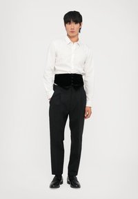 Weißes Hemd mit Knöpfen, schwarzer Samtweste, schwarze Hose und schwarze Schuhe. Das Outfit zeichnet sich durch ein tailliertes Design und glatte Texturen aus.