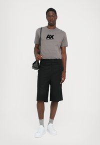 Pilka marškinėliai su "AX" logotipu, juodi šortai iki kelių, pilkos kojinės ir baltos sportbačiai. Modelis laiko juodą petinį kosmetinę.