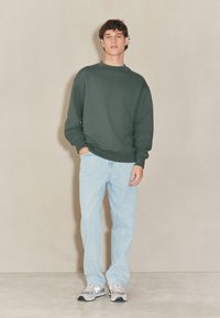 Grøn sweatshirt med rund hals og afslappet pasform, kombineret med lyseblå jeans. Modellen har grå og hvide sneakers på. Jævn tekstur over det hele.