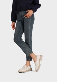 Scarpe slip-on bianche in pelle con superiore testurizzato e suola beije spessa, indossate con jeans scuri. Design semplice senza marchi visibili.