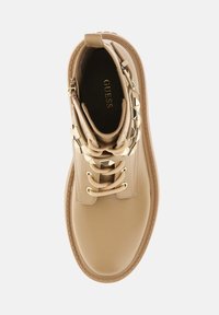Botín de tobillo beige con un acabado de cuero suave y brillante, detalles en hardware dorado y diseño de cordones. Presenta una puntera redonda y suela texturizada.