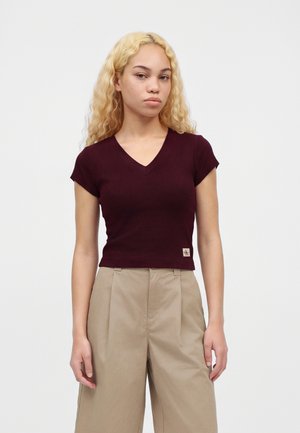 Jeune femme aux longs cheveux blonds bouclés, portant un haut bordeaux à manches courtes et col en V, ainsi qu’un pantalon beige taille haute, sur un fond uni.