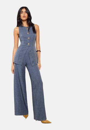 Vrouw staand, draagt een mouwloze blauwe op maat gemaakte jumpsuit met wijde pijpen, gouden knopen, gouden spitse schoenen en een bruine armband.