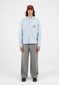 Gestreiftes blaues Hemd mit Knöpfen und einer Brusttasche, kombiniert mit grauen weitem Hosen und einer kastanienbraunen Mütze mit gelber Stickerei.