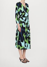 Šaty v zavinovacím stylu s výrazným abstraktním vzorem v modré, zelené a černé barvě. Dlouhé rukávy, výstřih ve tvaru V a odpovídající pásek zvýrazňují siluetu.