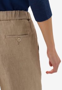 Pantalon en lin beige avec ceinture élastique, doté d'une poche arrière fermée par un bouton, présenté de profil avec un haut bleu.