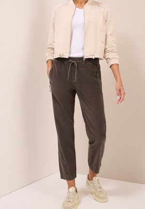 Beige Bomberjacke mit Reißverschluss, elastischen Bündchen; weißes Hemd darunter, dunkle Hose mit elastischem Bund und Kordelzug; helle Sneakers.