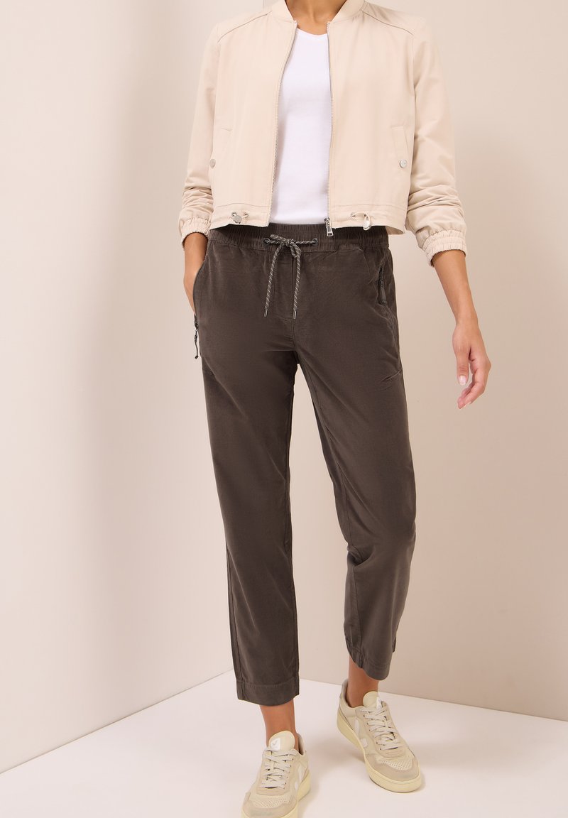 Veste bomber beige avec fermeture éclair, poignets élastiques ; chemise blanche en dessous, pantalon sombre avec taille élastique et cordon de serrage ; baskets claires.
