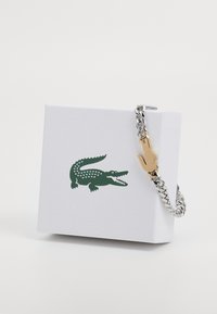 Bracelet en chaîne argentée avec un accent en crocodile doré, présenté sur une boîte blanche texturée arborant un logo crocodile vert.