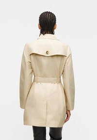 Άτομο με πλεκτά μαλλιά φοράει ένα μπεζ παλτό τρανς με ζώνη, βλέπουμε την πλάτη του, στέκεται μπροστά σε ένα απλό ανοιχτό γκρι φόντο.