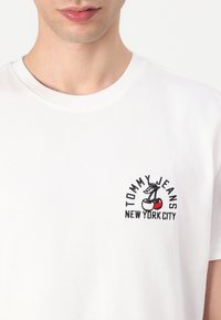White cotton t-shirt featuring a colorful embroidered design of cherries and text "TOMMY JEANS NEW YORK CITY" on the left chest.