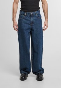 Wijde blauwe spijkerbroek met hoge taille, vijf zakken en metalen details. De textuur is glad met een vervaagde afwerking. Gecombineerd met zwarte schoenen.