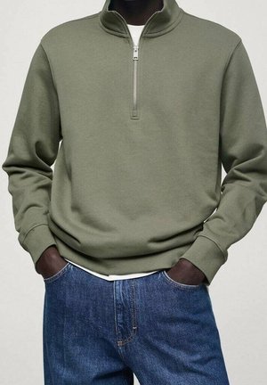 Dunkelhäutige Person trägt einen olivgrünen Pullover mit Viertelreißverschluss und hat die Hände in den Taschen einer blauen Jeans.