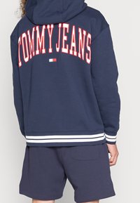 Marinblå hoodie med "TOMMY JEANS" i röda bokstäver på baksidan, med en vit randig nederkant och muddar, ihop med marinblå shorts.