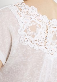 Blusa blanca con ribete de encaje que presenta un tejido texturizado y translúcido con un delicado patrón floral en el escote. Mangas cortas y corte relajado.