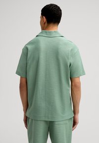 Camicia a manica corta in tessuto verde chiaro testurizzato. Presenta un colletto e un design dritto. Indossata con pantaloni coordinati.
