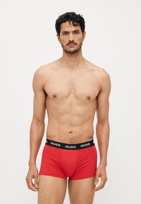 Boxer rouges en tissu doux avec une ceinture noire portant le logo blanc "HUGO". Le design est ajusté et lisse au toucher.