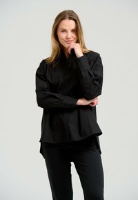 Sort skjorte med knapper lavet af glat stof, med krave, lange ærmer og en let buet kant. Båret med sorte bukser.