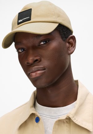 Mladý muž s béžovou čepicí s černou nášivkou loga a béžovou bundou přes bílé tričko na jednobarevném bílém pozadí.