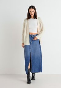 Cremefarbener Kabelstrick-Cardigan über einem weißen Cropped-Top, kombiniert mit einem blauen Denim-Maxirock mit einem Schlitz vorne und schwarzen Knöchelschuhen.