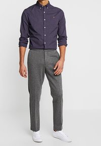 Camicia blu navy con bottoni, decorata con un piccolo logo sul petto, abbinata a pantaloni grigi slim-fit e sneakers bianche. Tessuto rigido e compatto con maniche lunghe.