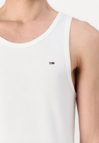 Fehér pamut ujjatlan felső, kerek nyakkivágással és vékony pántokkal, a mellrészen egy kis, piros, fehér és kék hímzett logóval.