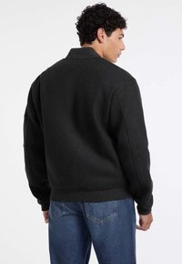 Giacca bomber nera in misto lana con colletto e polsini a coste. Presenta una vestibilità comoda con tasche laterali, abbinata a jeans in denim blu.