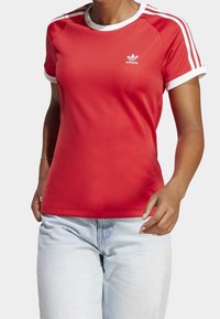 Czerwona sportowa koszulka z białymi akcentami i trzema paskami na ramionach. Gładki materiał, okrągły dekolt i logo Adidas na klatce piersiowej.