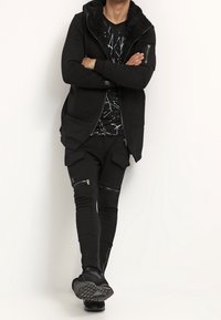 Felpa nera con zip e foderatura in pelliccia, abbinata a una t-shirt nera con grafica, pantaloni slim-fit con dettagli in zip e sneakers nere.