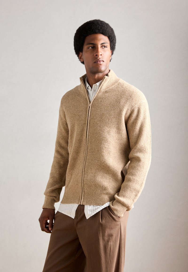 Beige, ribbes zip-up genser med høy krage, båret over en hvit og marineblå stripet skjorte, parret med brune bukser. Glatt tekstur.
