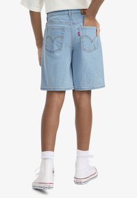 Άτομο που φοράει ανοιχτό μπλε τζιν σορτς και λευκά ψηλά αθλητικά Converse, στέκεται με το ένα χέρι στην πίσω τσέπη.