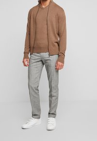 Mand iført brun zip-up sweater, matching brun skjorte, grå bukser og hvide sneakers står mod en grå baggrund.