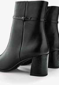 Paire de bottines noires en cuir avec talons carrés et une fine sangle ornée d’un petit détail en forme de croix autour de la partie supérieure.