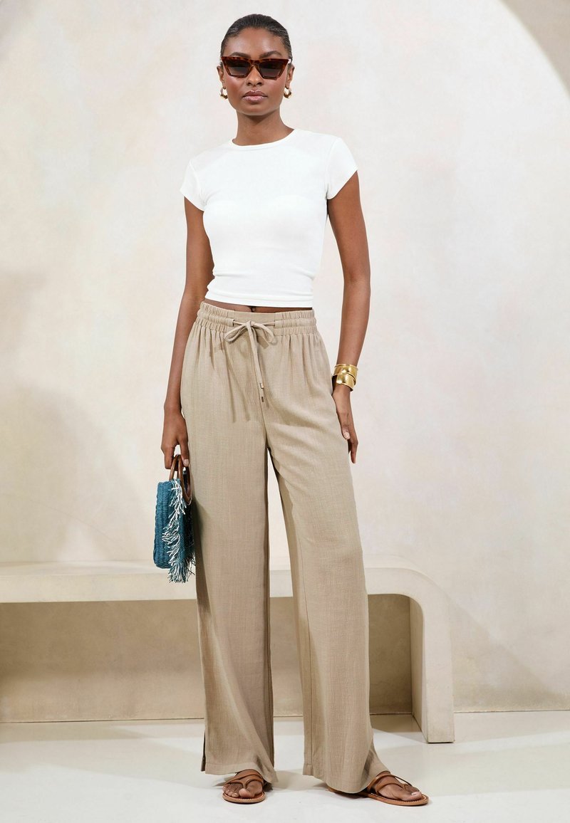 Nő teknőspáncél mintás napszemüveget visel, fehér rövidített pólót, bézs bő szárú nadrágot, barna szandált, arany ékszereket, kéket rojtos kézitáskát tartva.