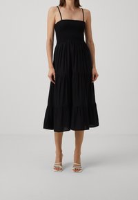 Femme portant une robe midi noire sans manches froncée avec une jupe à volants et des sandales blanches à talons hauts à brides sur un fond neutre.