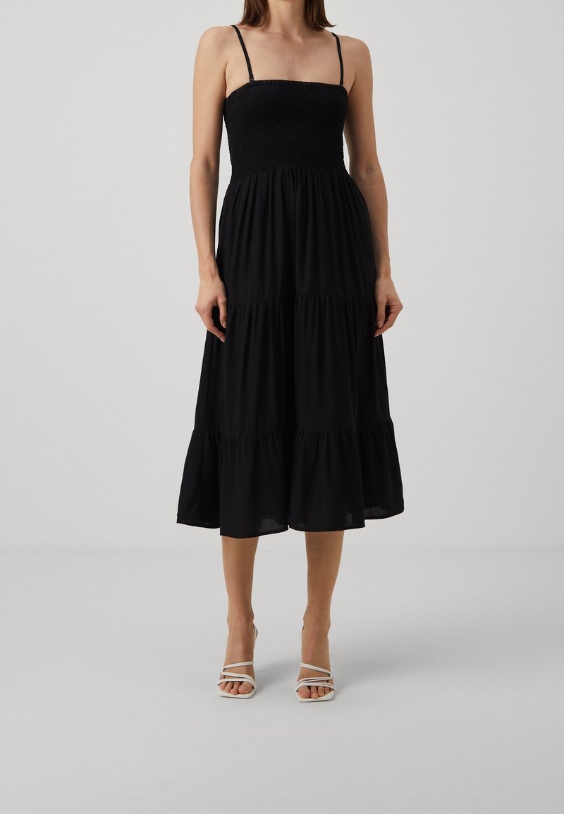 Femme portant une robe midi noire sans manches froncée avec une jupe à volants et des sandales blanches à talons hauts à brides sur un fond neutre.