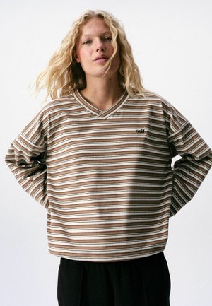 Blonde woman with wavy hair wears a beige and brown striped long-sleeve top with small "self:" text on chest, standing against a plain background.