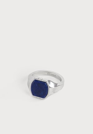 Anneau en argent avec une pierre hexagonale bleue posée dessus et le mot "GUESS" gravé à l'intérieur de l'anneau.