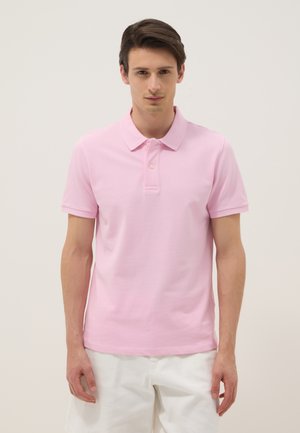 Jonge man staat recht naar voren kijkend, draagt een lichtroze polo met korte mouwen en een witte broek tegen een effen achtergrond.