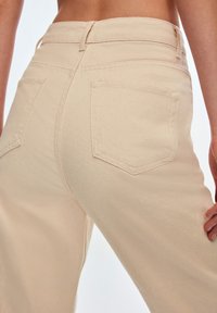 Vaqueros de tiro alto beige con pierna recta, que cuentan con dos bolsillos traseros y costuras visibles, elaborados con tela vaquera de algodón.