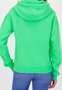 Persona che indossa una felpa con cappuccio verde brillante e jeans blu, mostrata di spalle su uno sfondo bianco semplice.